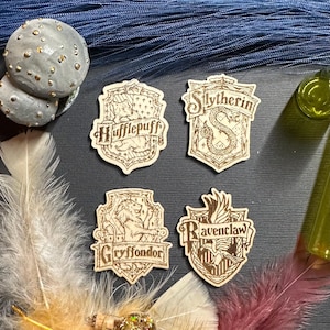 May include: Four wooden Harry Potter house crests: Hufflepuff, Slytherin, Gryffindor, and Ravenclaw. Each crest is intricately etched with house symbols and names. The crests are displayed with feathers, a small bottle, and other decorative elements.