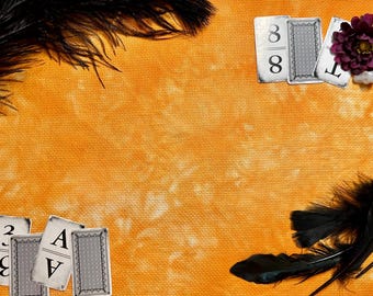 May include: A textured orange fabric background featuring black feathers in the top left and bottom right corners. Playing cards are arranged in the corners, with a dark purple flower and small white stones placed near the top right cards.