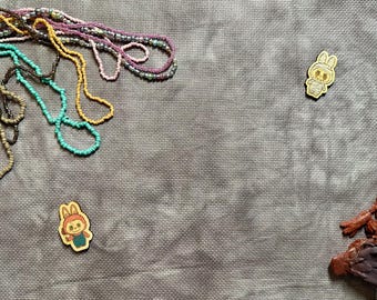May include: Several strands of colorful beaded necklaces in shades of pink, yellow, turquoise, and brown, two small wooden character charms, and a dark brown textured object are arranged on a gray fabric surface with a subtle grid pattern.