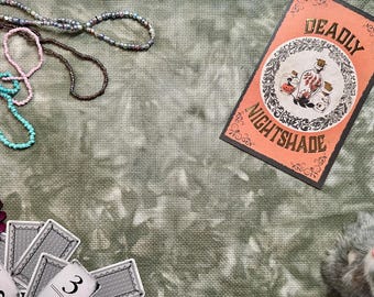 May include: A mottled olive green fabric background featuring a rectangular card labeled DEADLY NIGHTSHADE with a potion bottle illustration, strands of multi-colored beads, a dark purple flower, several playing cards, and a small gray plush animal.