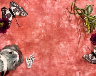 May include: A coral-colored fabric with a tie-dye pattern featuring two decorative butterflies, two dark purple flowers, a small air plant, and a miniature white hand figurine with black palmistry lines printed on the surface of the fabric.