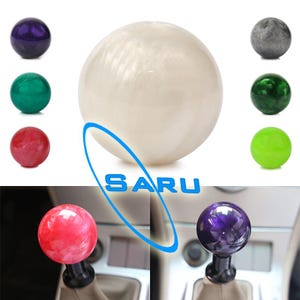Op de afbeelding: Een verzameling kleurrijke, bolvormige versnellingspookknoppen. De knoppen worden getoond in verschillende kleuren, waaronder wit, paars, groen, rood en grijs. Het woord "SARU" wordt in blauw weergegeven. Twee knoppen worden in een voertuig getoond.