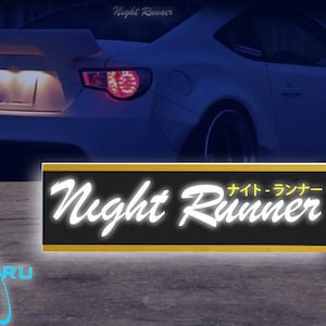 Pode incluir: Um autocolante retangular preto e dourado com as palavras "Night Runner" em uma fonte branca estilizada. Caracteres japoneses estão acima da palavra "Runner". Um carro esportivo branco ao fundo.
