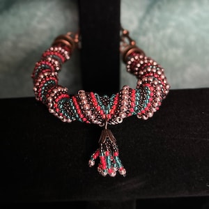 May include: A beaded bracelet with a spiral design in shades of pink, teal, and brown. The bracelet has a tassel made of the same beads.