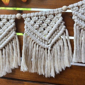 May include: A white macrame banner with fringe hanging on a wooden surface. The banner is made of knotted string and has wooden beads strung between each section.