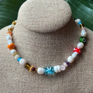 May include: A white pearl necklace with colorful beads, including a blue evil eye charm, a red mushroom charm, and orange and brown beads.