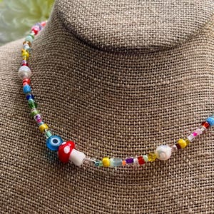 Puede incluir: Un collar de cuentas de colores con un dije de hongo y una cuenta de ojo turco. El collar presenta una variedad de pequeñas cuentas redondas en múltiples colores, incluyendo rojo, azul, amarillo y verde. El collar se exhibe sobre una superficie de tono neutro.