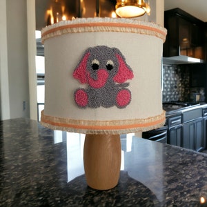 May include: A lampshade with a light brown fabric base and a light orange trim. The lampshade features a grey embroidered elephant with pink ears and a pink trunk.