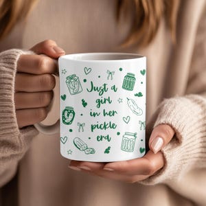May include: A white ceramic mug featuring green line art illustrations of pickle jars, individual pickles, hearts, stars, and bows. The text in the centre reads, "Just a girl in her pickle era," printed in a dark green, handwritten-style font.