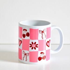 May include: White ceramic mug with a pink and red checkered pattern. The pattern includes cherries, hearts, bows, and a flower with a smiley face.