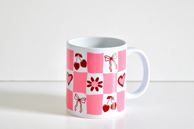 May include: White ceramic mug with a pink and red checkered pattern. The pattern includes cherries, hearts, bows, and a flower with a smiley face.