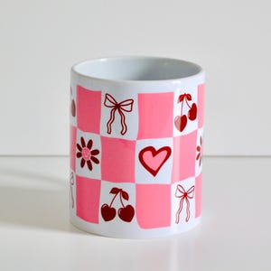 May include: A white ceramic mug with a pink and white checkered pattern. The mug has red illustrations of hearts, bows, and cherries.