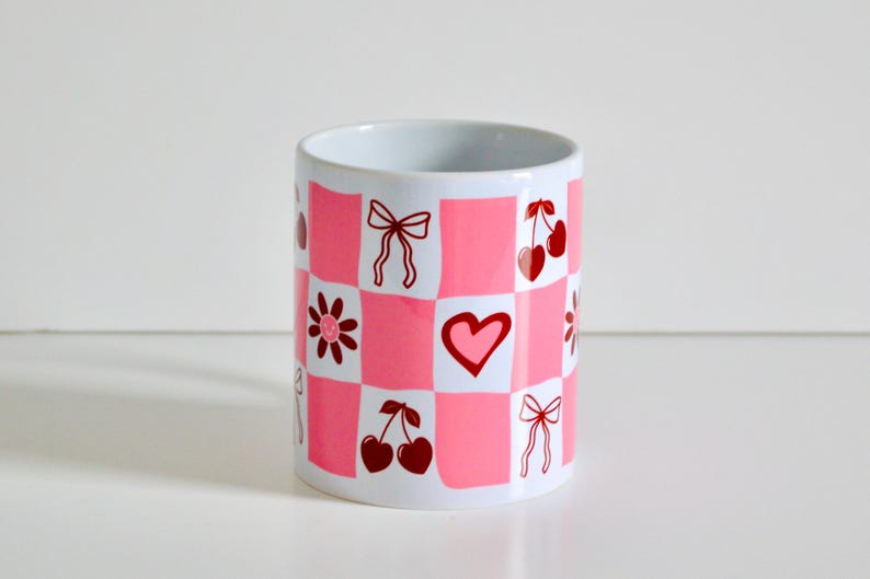 May include: A white ceramic mug with a pink and white checkered pattern. The mug has red illustrations of hearts, bows, and cherries.