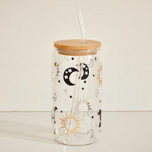 May include: A clear glass reusable tumbler with a bamboo lid and two glass straws. The tumbler features a celestial design with black moons, stars, and gold suns.