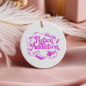 May include: A round white ceramic ornament featuring the purple text Fiction Addiction in a stylized font, surrounded by illustrations of open and closed books. The ornament is suspended by a thin gold-colored string at the top center.