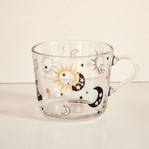 May include: A clear glass mug with a handle, decorated with celestial designs. The mug features gold, black, and white suns, moons, and stars. The mug is designed for hot or cold beverages.