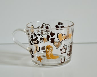 Peut inclure: Une tasse en verre transparent avec une anse présente un motif sur le thème du western. La tasse est décorée de bottes de cowboy brunes, noires et dorées, de fers à cheval, d'étoiles et de motifs de peau de vache.