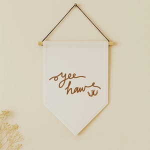 May include: A white banner with the words "yee haw" in brown script, along with two brown horseshoe illustrations. The banner is suspended from a wooden dowel and black string.