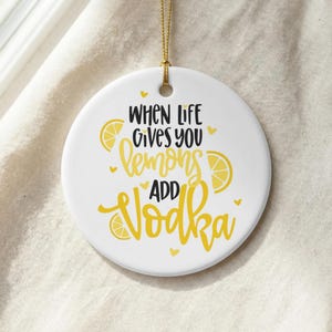 May include: A round white ceramic ornament hanging from a gold string. The ornament features the black and yellow text "WHEN LIFE GIVES YOU lemons ADD Vodka" with yellow lemon slice illustrations and small yellow hearts scattered around the text.