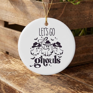 May include: A round white ceramic ornament featuring a black graphic of three ghosts wearing cowboy hats. The central ghost holds a lasso. The text "LET'S GO" appears above the ghosts, and the word "ghouls" is written below in a stylized font.