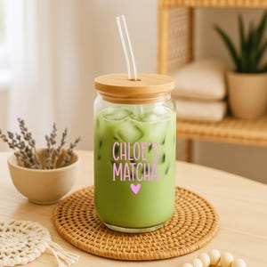 May include: A clear glass tumbler with a bamboo lid and a glass straw, filled with a green matcha beverage and ice cubes. The glass features the pink text "CHLOE'S MATCHA" followed by a small pink heart graphic printed on the front surface.