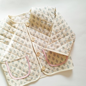 May include: A cream-colored quilted jacket with a floral print and pink trim. The jacket has a button closure and two patch pockets.