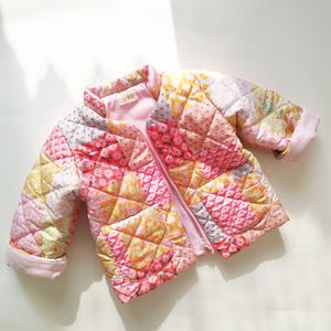 May include: A quilted jacket in a patchwork design, featuring a mix of pink, yellow, and peach fabrics. The jacket has a full zipper, a small pocket, and a gingham lining. The sleeves have a rolled cuff. The jacket is designed for a child.
