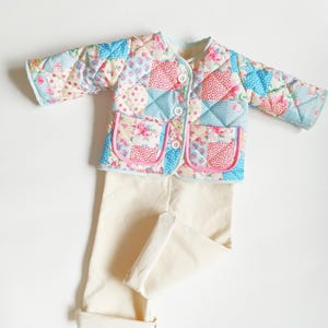 Puede incluir: Conjunto de chaqueta y pantalón acolchados para bebé. La chaqueta presenta un diseño de patchwork con estampados florales y geométricos en rosa, azul y crema. La chaqueta tiene dos bolsillos con ribete rosa y botones blancos. El pantalón es de color crema.