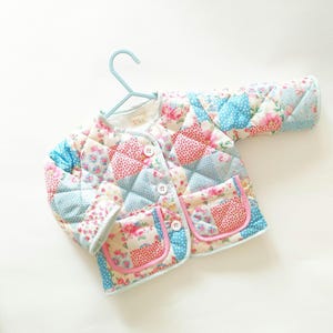 May include: A quilted jacket featuring a patchwork design of floral and polka dot patterns in shades of pink, blue, and white. The jacket has long sleeves, a round neckline, three white buttons, and two front pockets with pink trim. A label reads Pia.