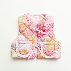 Könnte beinhalten: Eine gesteppte Weste mit einem Patchwork-Design aus verschiedenen Blumen- und geometrischen Mustern in Rosa-, Gelb- und Weißtönen. Die Weste hat zwei Vordertaschen mit gerüschten Kanten, einen Vichy-Besatz und zwei Bindebänder. Ein Etikett mit der Aufschrift Pia ist sichtbar.