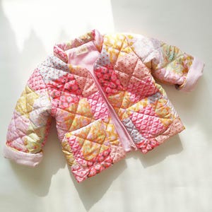 May include: A quilted jacket featuring a patchwork design of various floral and geometric patterns in shades of pink, yellow, and white. The jacket has a front zipper closure, a stand-up collar, two front pockets, and light pink gingham cuffs.
