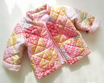 May include: A quilted jacket featuring a patchwork design of various floral and geometric patterns in shades of pink, yellow, and white. The jacket has a front zip closure, a stand-up collar, two front pockets, and light pink gingham cuffs.