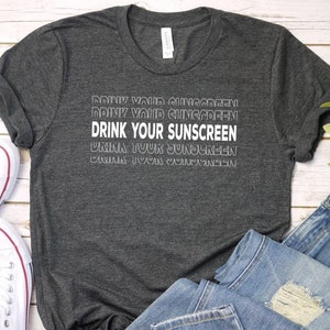 May include: A dark gray t-shirt with the text "DRINK YOUR SUNSCREEN" in white, with the same text repeated in a faded outline. The shirt is paired with light blue jeans and white canvas sneakers.