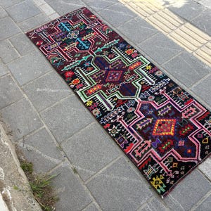 May include: A rectangular, patterned rug with a dark burgundy base and intricate geometric designs in various colors, including red, green, blue, and yellow. The rug is placed on a gray stone surface, showcasing its detailed craftsmanship.