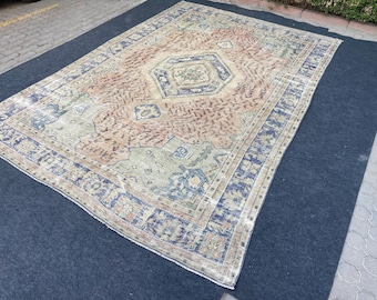 May include: A large, rectangular, vintage rug with a central medallion design. The rug features a faded color palette of peach, blue, and beige. The border is a darker blue with floral patterns. The rug is placed on a dark blue surface.