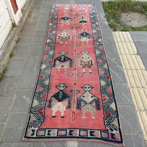 May include: A long, rectangular rug with a pink background and a repeating pattern of stylized human figures. The rug has a border of blue, red, and white geometric designs. The rug is likely made of woven fabric.