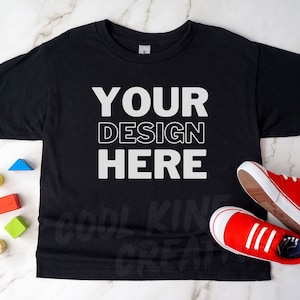 May include: Black t-shirt with the text "YOUR DESIGN HERE" in white. Red trainers with white soles and blue and white striped socks are next to the shirt. Colourful wooden blocks are scattered around the shirt.