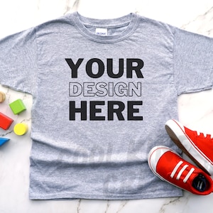 May include: A gray t-shirt with the text "YOUR DESIGN HERE" printed on the front. The shirt is lying flat on a white marble surface with colorful wooden blocks and red sneakers.