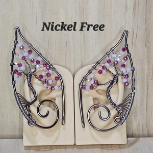 May include: A pair of silver wire ear cuffs with pink and white beads, shaped like a tree branch. The text "Nickel Free" is visible above the cuffs.