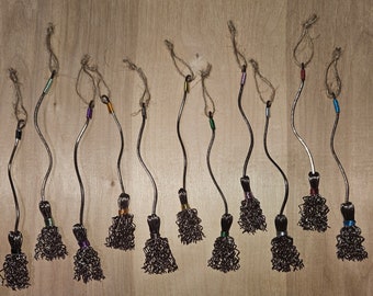 May include: A collection of 14 miniature brooms made of wire and twine, with colorful beads at the top. The brooms are hanging from a wooden surface.