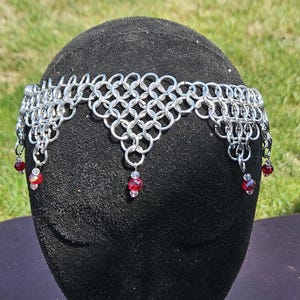 May include: A silver chainmail crown with a pointed top and dangling red crystal beads.
