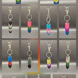 May include: A collection of keychains with various pride flag designs. Each keychain features a series of heart-shaped metal scales in different colors, attached to a silver-colored ring and clasp. The color combinations represent different LGBTQ+ identities.