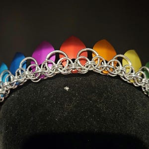 May include: A crown constructed from silver-toned metal jump rings woven in a chainmail pattern, featuring a series of pointed, petal-shaped scales in a vibrant rainbow gradient of blue, purple, red, orange, yellow, and green colors.
