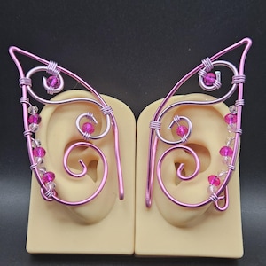 Op de afbeelding: Een paar roze draad elf oormanchetten met decoratieve roze en heldere kralen. De draad is gevormd in een oorachtig ontwerp, met een gebogen spiraaldetail. De manchetten worden gepresenteerd op een lichtbeige oormodel tegen een zwarte achtergrond.