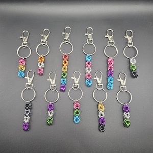May include: A collection of handmade keychains with metal clasps and rings. Each keychain features a chainmail-style design in various colors, including rainbow, pink, blue, and green. The keychains are displayed against a dark background.