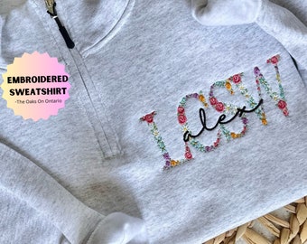 May include: A gray sweatshirt with the name "Alex" embroidered in a floral design. The sweatshirt has a zipper and is made of a soft, comfortable fabric.