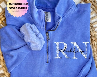 May include: A blue embroidered sweatshirt with a white floral design that spells out "RN" and the name "Ashley".