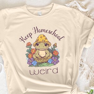 May include: Cream-colored t-shirt with the text "Keep Homeschool Weird" and a cartoon frog wearing a mushroom hat. The frog is surrounded by colorful flowers. The shirt is knotted at the bottom.