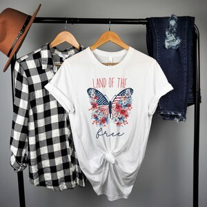 May include: White t-shirt featuring a red, white and blue butterfly graphic. The butterfly has the American flag on its wings and is surrounded by red, white and blue flowers. The text "Land of the free" is printed below the butterfly.