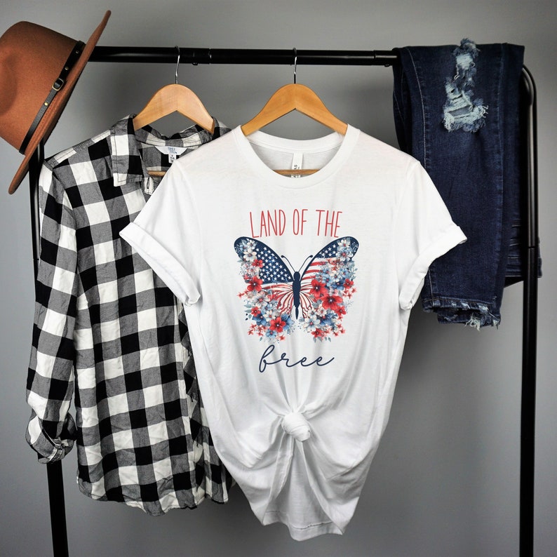 May include: White t-shirt featuring a red, white and blue butterfly graphic. The butterfly has the American flag on its wings and is surrounded by red, white and blue flowers. The text "Land of the free" is printed below the butterfly.